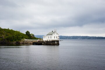 Historic white wooden building perched on a rocky shore overlooking a calm lake surrounded by lush, forested hills on a cloudy day.