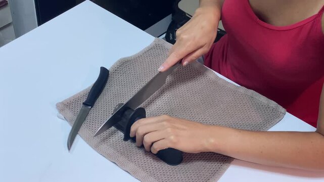 A woman carefully sharpens a kitchen knife using a black sharpener on a brown towel, ensuring a keen edge for her culinary tasks. Another knife rests beside the sharpener