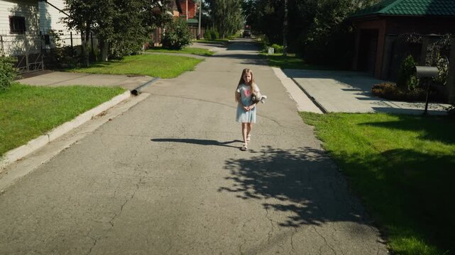 Fair skin girl squinting under bright sun walks alone on cracked suburban street with hands locked together and stuffed toy in arm, mailbox visible on side near residential homes and green lawn