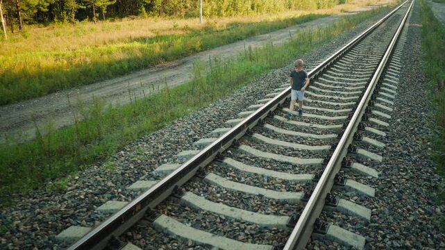 young boy dressed in casual clothes walking alone along rail track in rural setting, passing sign post beside railway with forest and dirt path in background during quiet sunny afternoon