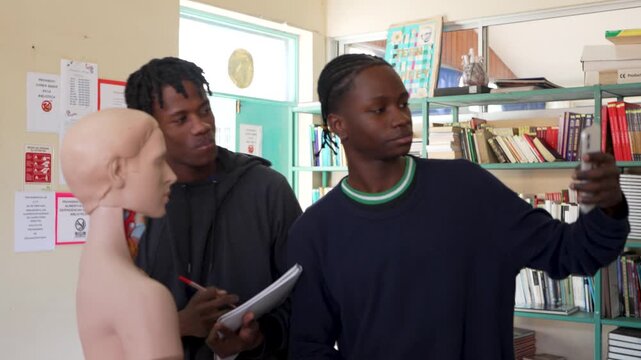Two high school students using a phone and notepad while studying a human anatomy mannequin in class