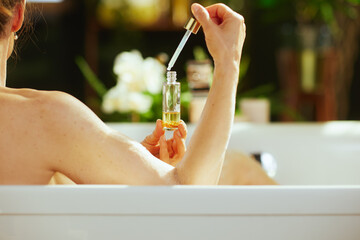 Fototapeta premium A woman, seen from behind, dispenses yellow bath liquid via dropper into a white bathtub. Serene blurred green foliage & white flowers enhance the mood of tranquility & personal care.