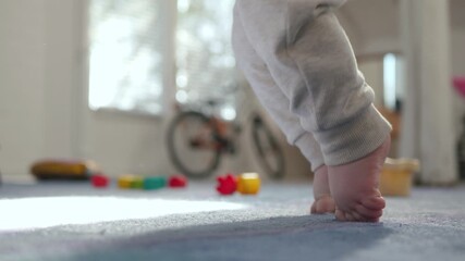 In a bright, cozy living space, unrecognizable little baby learns and practices standing barefoot on a soft carpet, surrounded by colorful toys and a brightly colored bicycle - Powered by Adobe