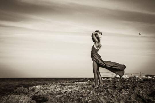 Beautiful lonely woman standing on sea shore in elegant fluttering dress and wind in her hair and looking into the horizon of sea in black and white dramatic colors with flying lonely seagull in the s