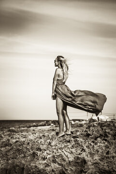 Beautiful woman standing on sea shore in elegant fluttering dress and wind in her hair and looking into the horizon of sea in black and white dramatic colors 