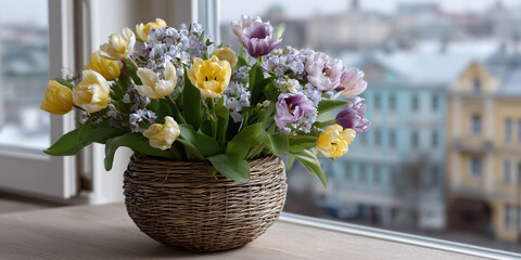Colorful spring flowers in a woven basket on a window sill with a city view in the background