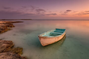Obraz premium A serene sunset over a calm ocean with a small boat resting in shallow water, reflecting the pastel sky and soft clouds, evoking tranquility and peace.