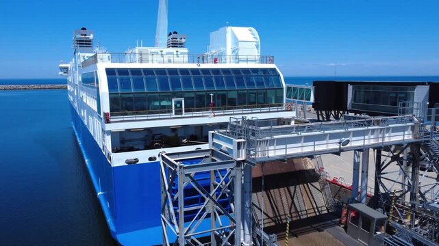 The aft deck of an elegant blue ferry moored at a dock and its boarding structure, with its glass decks and raised boarding bridge in a blue decor. Matane, Quebec, Canada, 2025.
