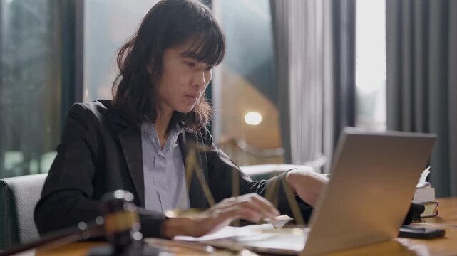 Professional woman in law office working on laptop with legal documents, justice scale, and gavel.