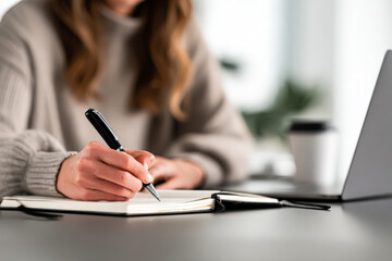Female individual is engaged in writing notes in a notebook at a stylish workspace, with a laptop and coffee cup nearby, reflecting a productive atmosphere