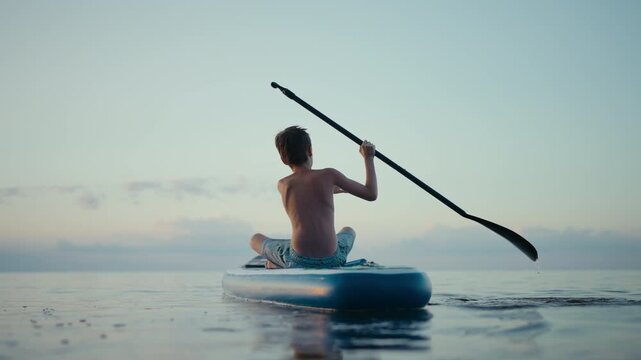 A boy rows while sitting on a SUP board at sunset
