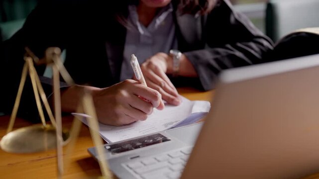 Focused legal expert woman taking notes from law book, justice scale and gavel visible on table.