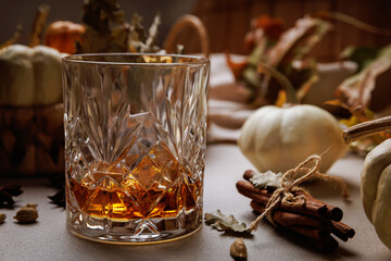 Cut glass filled with whiskey surrounded by cinnamon sticks, cardamom and pumpkins on soft fabric..