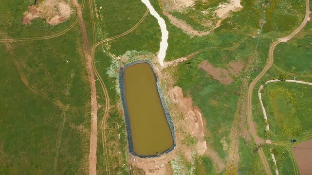 Drone shot shows agricultural water reservoir construction. A newly lined irrigation pond in a green field stores water for farm use.  Workers install protective geomembrane liner over compacted base.