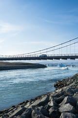Scenic view of a modern suspension bridge spanning a tranquil river, with rocky shoreline and icebergs floating in the water, showcasing natural beauty and engineering marvel