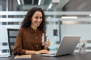 Smiling woman engaged in a virtual conference or online meeting, holding a glass of water, while...