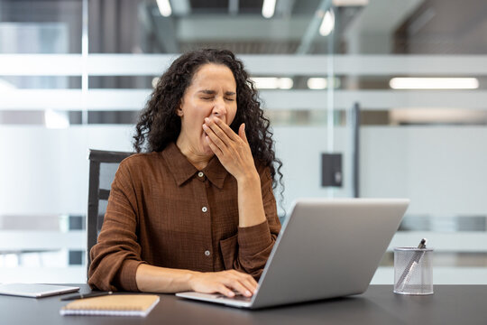 Businesswoman yawning and covering her mouth at a modern office desk, tired and overworked while working on a laptop, showing burnout, fatigue and low energy during long hours
