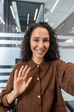 Fototapeta Young woman with curly hair smiling and waving during a virtual meeting or online conference, communicating remotely from a modern business office environment