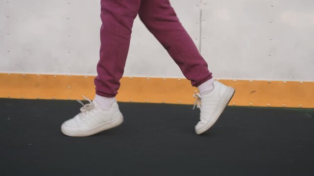 Female coach jogging on curved asphalt court beside barrier wall with yellow edge, close up of legs clad in purple tracksuit and white sneakers stepping on sports surface under soft pastel dusk light
