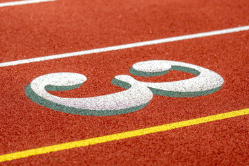 Close up photo of outdoor running track lane white number three, 3, with green shadow, on a new red track with white lane lines.