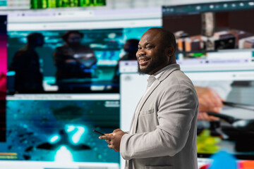 Cheerful man browses social media feed on smartphone in front of digital video wall showcasing multimedia content. Happy viewer in modern space surrounded by diverse multimedia clips.