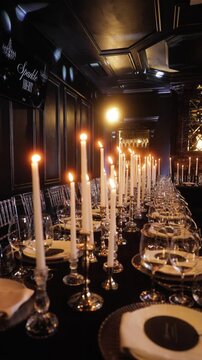 Luxury banquet table with tall white candles, elegant glassware and plates in dark room, warm candlelight reflections creating romantic evening atmosphere for wedding reception or gala dinner