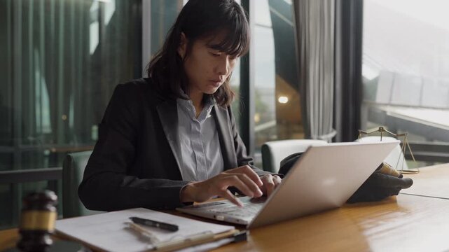 Modern female lawyer using laptop at office desk with justice scale, gavel, and legal documents.