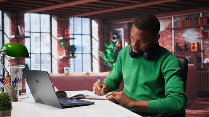 African american male teleworker writing personal notes at his desk in modern loft, remote job....