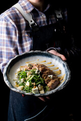 A skilled chef presents a beautifully plated dish featuring tender meat, vibrant greens, and crunchy nuts. The setting, dimly lit, adds warmth to the culinary creation