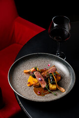 A beautifully arranged plate holds tender meat and colorful vegetables, bursting with flavors. A glass of red wine sits on a dark table beside the dish, enhancing the inviting atmosphere