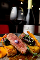 A beautifully plated steak rests on roasted vegetables, garnished with petals. Behind, a glass of red wine and a bottle add elegance to the cozy dining atmosphere