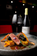 A beautifully plated gourmet dish showcases tender meat paired with vibrant vegetables, set against a backdrop of soft lighting. A glass of red wine completes the elegant setting