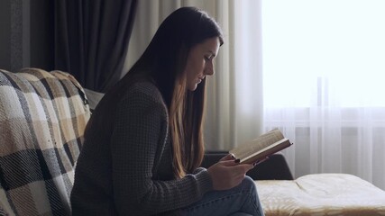 A young woman sitting on a comfortable sofa in a cozy living room, reading a book and relaxing at home. Concept of peaceful leisure, weekend rest, and self-care moments. Warm soft light creates a calm - Powered by Adobe