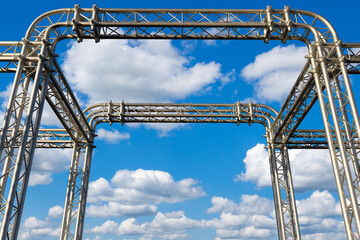 Construction, Music, Entertainment. Metallic stage trusses and rigging system forming structural support for concert lighting and sound equipment against blue sky.