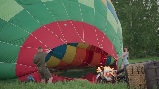 couple hold hot air balloon envelope open while burner fan heats air into vibrant checker patterned fabric for inflation during calm sunset pre flight preparation in serene grassy field