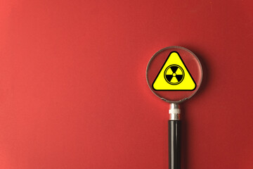 Radiation hazard identification and safety concept. A magnifying glass focuses on a radioactive warning sign on a red background, symbolizing the inspection and assessment of nuclear or toxic risks.