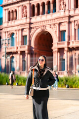 Fototapeta premium Happy young Latina woman wearing a black dress, white shearling jacket and sunglasses, smiling in front of Casa Rosada, Argentina’s presidential palace located in Plaza de Mayo, Buenos Aires