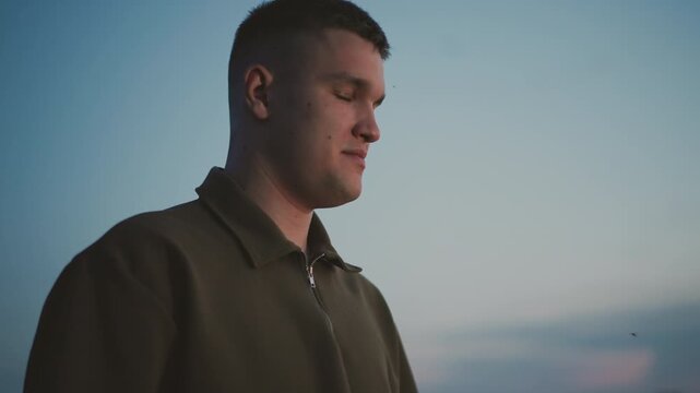 close up of young man standing in grassy field at dusk with insects buzzing around his head under clear sky capturing contemplative expression and tranquil outdoor moment as twilight settles
