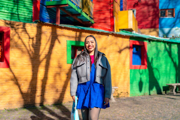 Obraz premium Happy young Latina woman in a bright blue dress and gray jacket smiling in Caminito, Buenos Aires, surrounded by vibrant buildings and rich cultural street art atmosphere, Argentina
