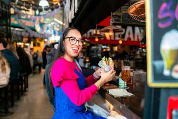 Young happy Latina enjoying choripan and craft beer at Mercado de San Telmo, a top foodie spot in Buenos Aires for authentic street food and local gastronomy