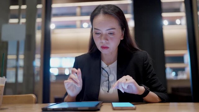 Focused Asian woman multitasking with tablet and smartphone in cafe. Smart professional in black blazer working efficiently with digital tools.
