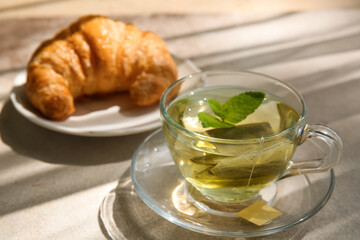 Cup of hot tea with mint leaves and croissant on light background