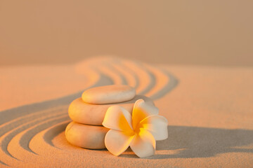 Stack of spa stones with plumeria flower on light sand at sunset. Zen concept