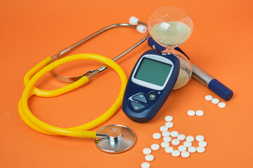 pills, stethoscope and digital blood glucose meter on orange background, hourglass as a symbol of...