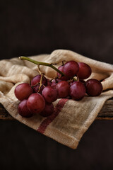Red Grape Still Life. Closeup of  a bunch of ripe red grapes on a kitchen towel draped over the edge of a rustic wooden table with a mottled brown background.