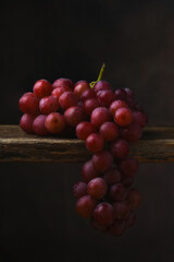 Red Grape Still Life. A bunch of ripe red grapes draped over the edge of a rustic wooden table with a mottled brown background.