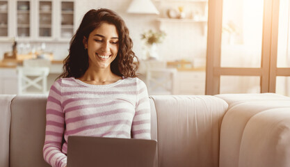 A young woman sits on a comfortable sofa using a laptop. She is smiling and appears engaged with her work. The sun is shining through the window, creating a warm atmosphere.