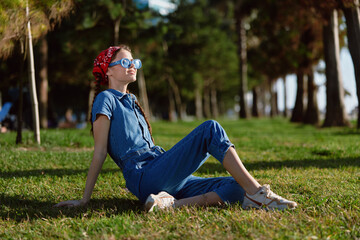 Fototapeta premium lifestyle woman in modern boho-western denim street style relaxes outdoors on grass with film color effect, wearing sunglasses and red headscarf on sunny day