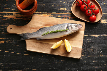 Cutting board with fresh raw seabass fish, lemon and cherry tomatoes on black wooden background