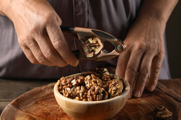 Elderly woman cracking walnuts with nutcracker into bowl on wooden table
